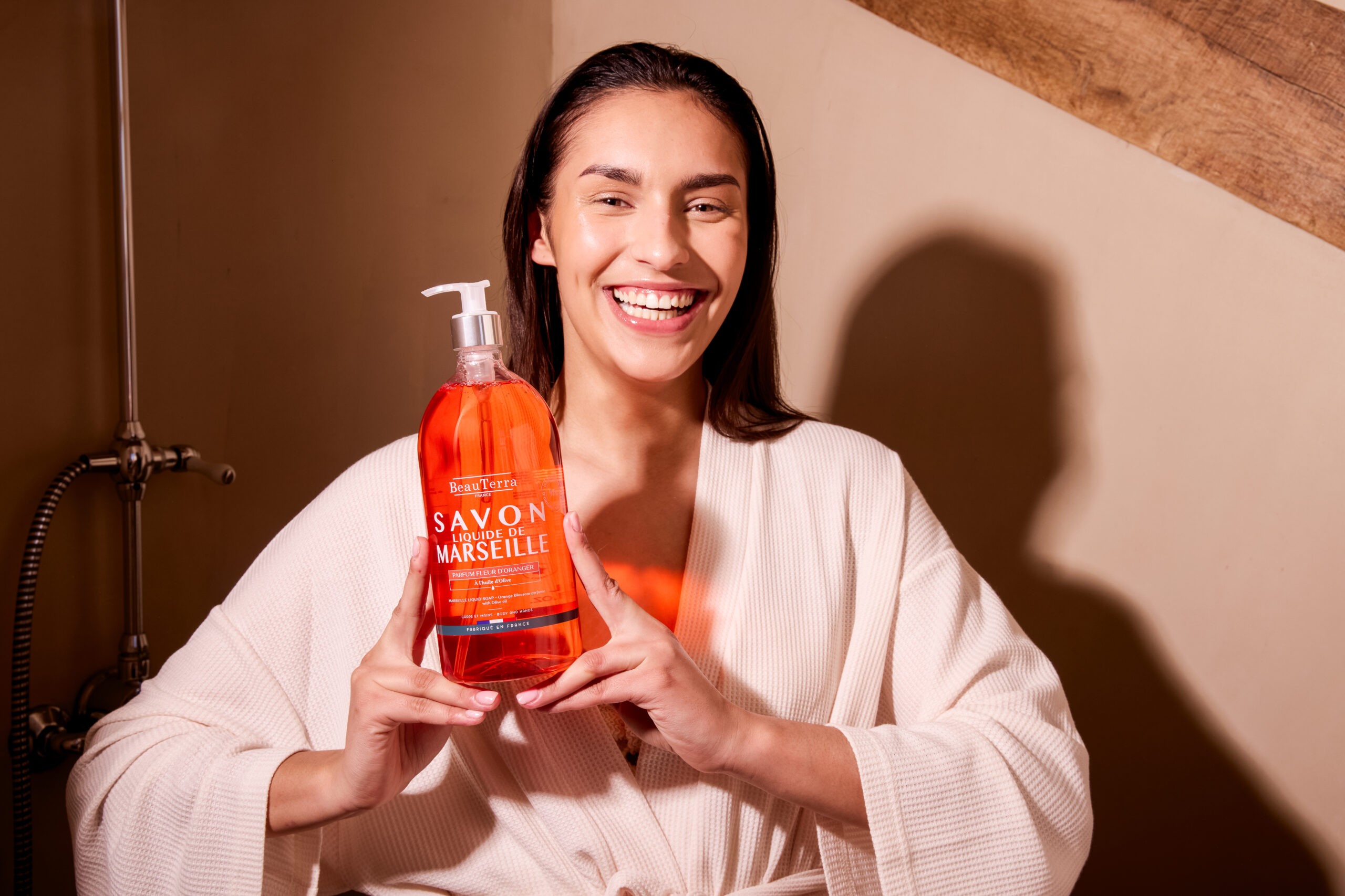 Une femme souriante tient un Savon Liquide de Marseille BeauTerra Fleur d'Oranger dans une salle de bain lumineuse.
